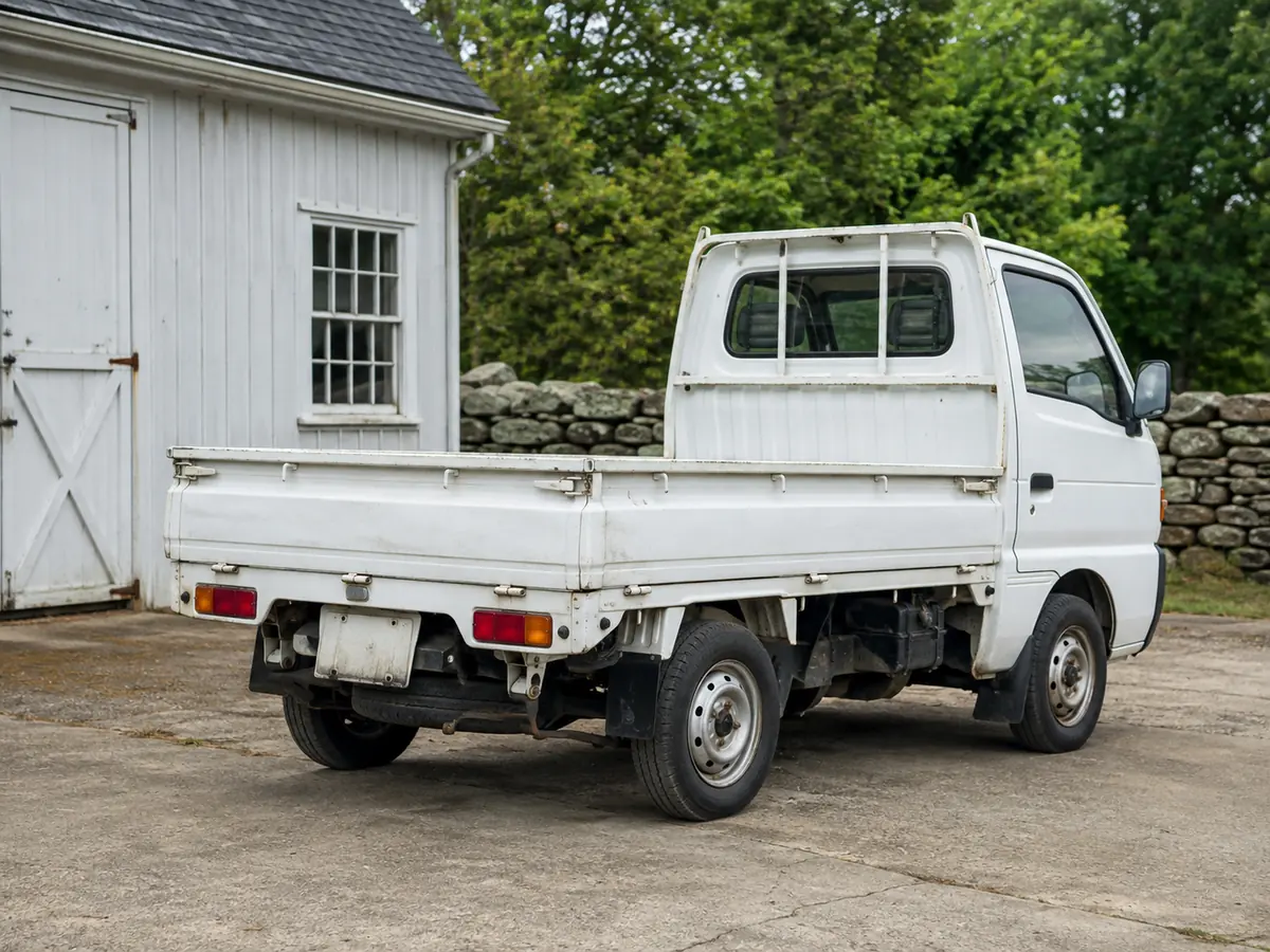 Rear three-quarter view of a white kei pickup: bed, wheels and underside by a workshop